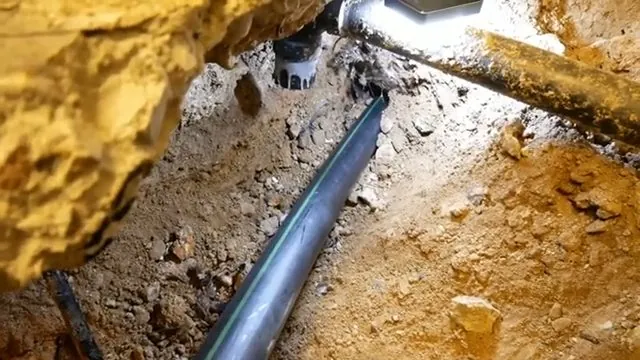 A black pipe lying in an excavated trench with an excavator bucket above the soil