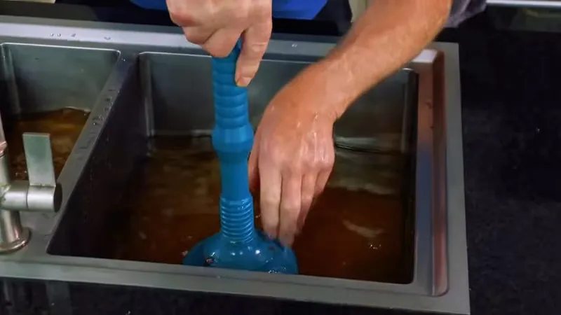 Hands holding a blue plunger-style tool over a stainless sink full of murky water