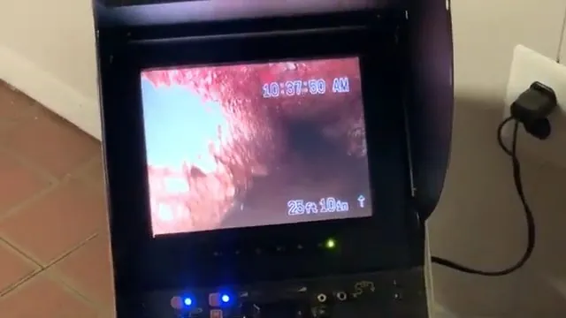 A camera inspection monitor on a tiled floor showing a reddish pipe interior
