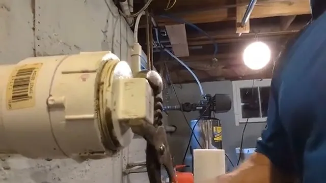 A hand using channel locks to loosen a cleanout cap in a basement, with a person standing beside