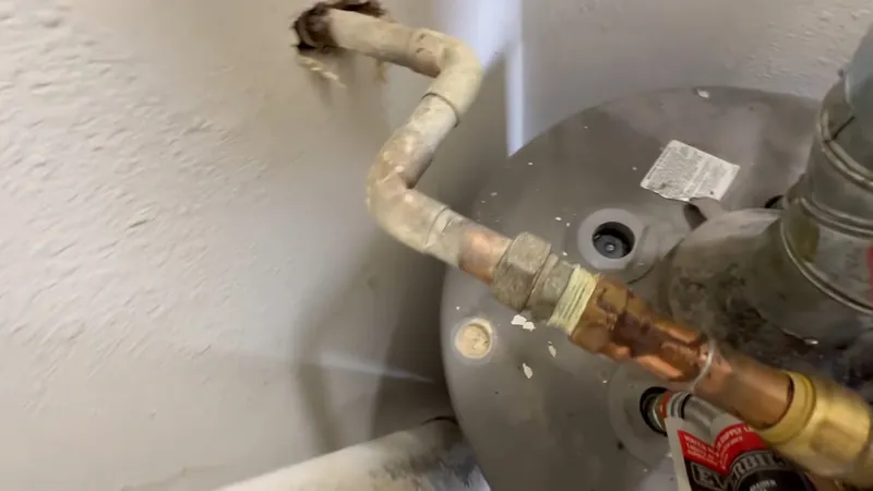 Corroded brass and copper fittings above a gray water heater tank against a white wall