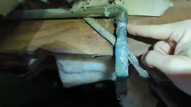 A hand touching a green corroded copper pipe elbow under a floor with insulation around it
