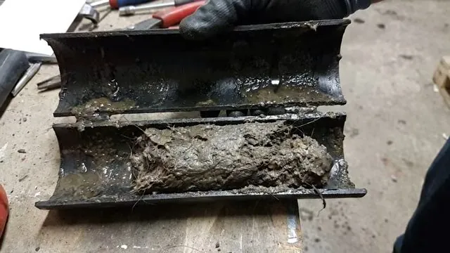 A black pipe cut open lengthwise on a workbench, its interior filled with gray sludge