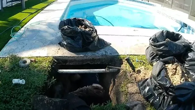 An excavated trench beside a swimming pool with a pipe exposed and black bags of soil on the grass
