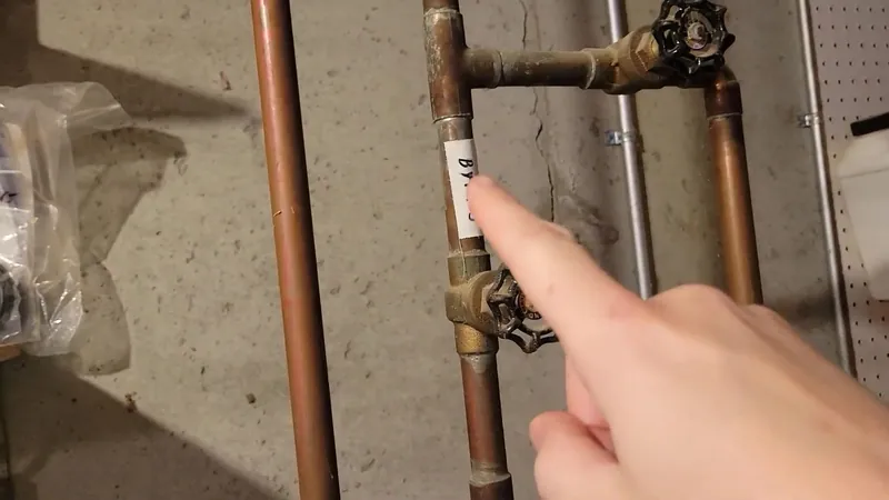 A finger pointing at a brass shutoff valve on copper pipes against a wall