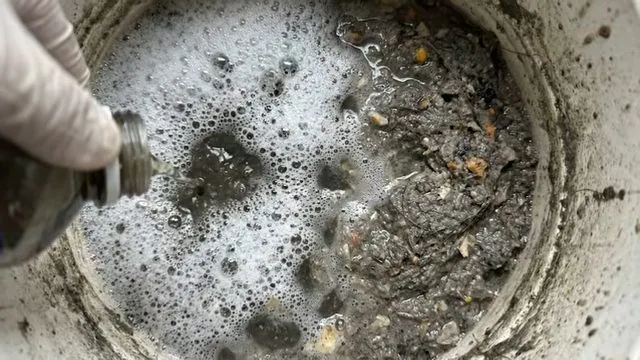 A gloved hand over a round drain filled with foamy dirty water
