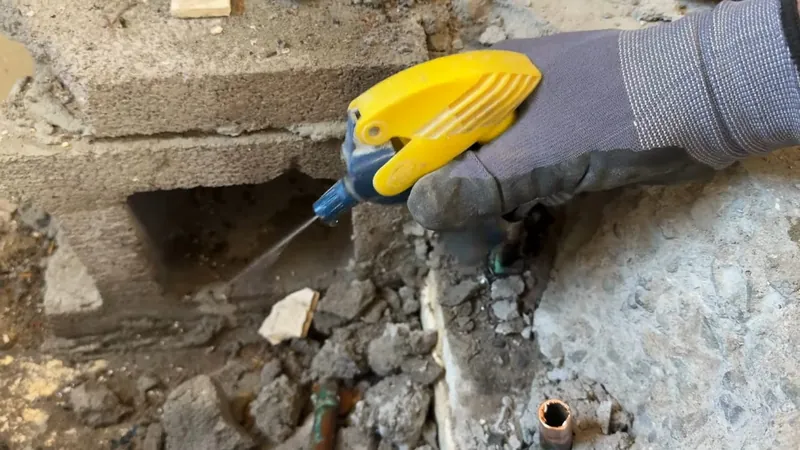 A gloved hand using a yellow tool at a hole broken into a concrete floor, a small copper pipe exposed below