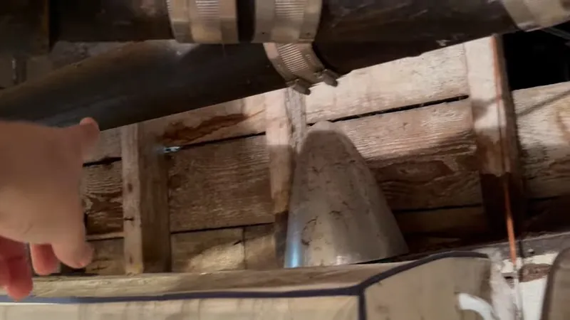A hand beside an old cast iron drain pipe running under wooden floor joists