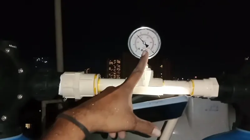 A hand beside a pressure gauge on white pipe fittings with city lights in the dark behind