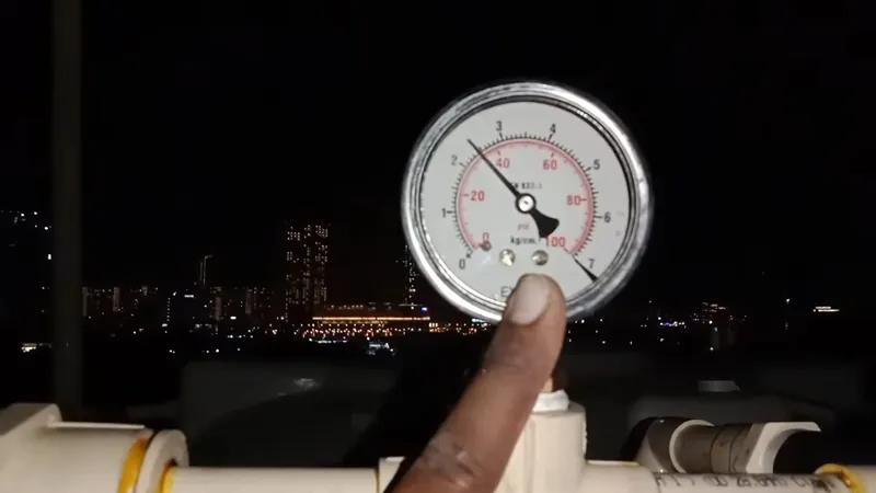 A hand pointing at a large round pressure gauge mounted on white pipes, with city lights in the dark behind