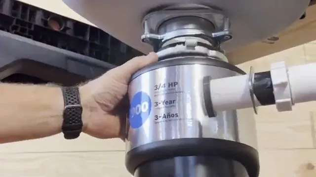 A hand holding a garbage disposal beside the white drain pipe under a counter