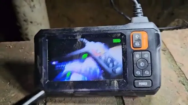 A handheld inspection camera monitor resting on a ledge, its screen showing a blue pipe interior