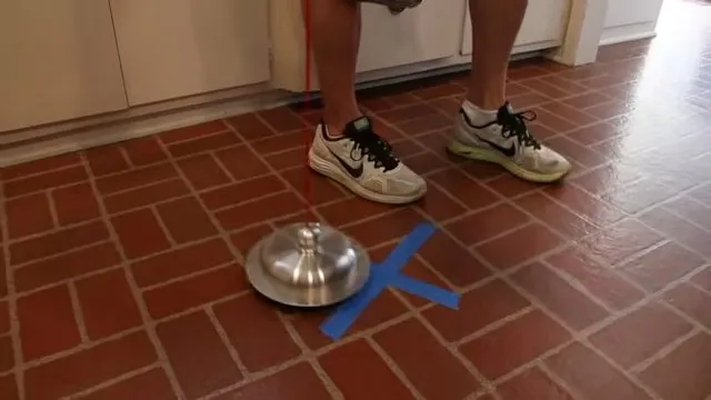 A person's legs beside a listening device on a red tile floor with a blue tape X marking a spot