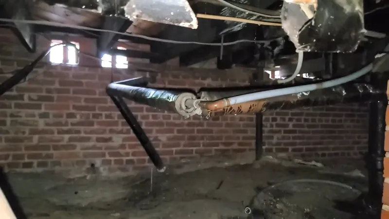 Old black and white pipes running under floor joists in a brick-walled crawlspace