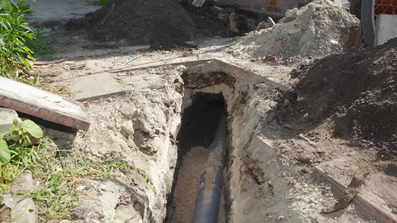 An open narrow trench beside a house with black pipe exposed and mounds of soil on either side