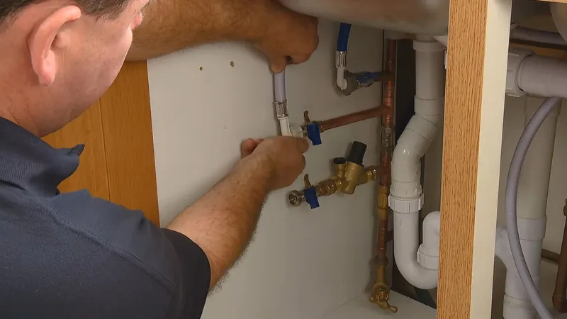A man connecting valves and supply lines under a sink beside the white drain pipes