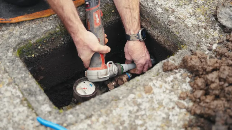 Hands using a power tool on a pipe inside a stone-lined meter box set in the ground