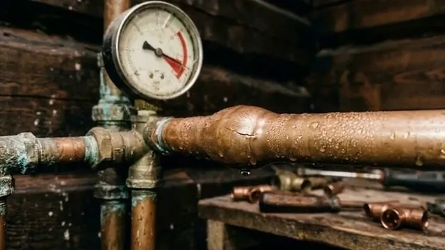 A pressure gauge mounted on a copper pipe manifold in a dark wood-framed space