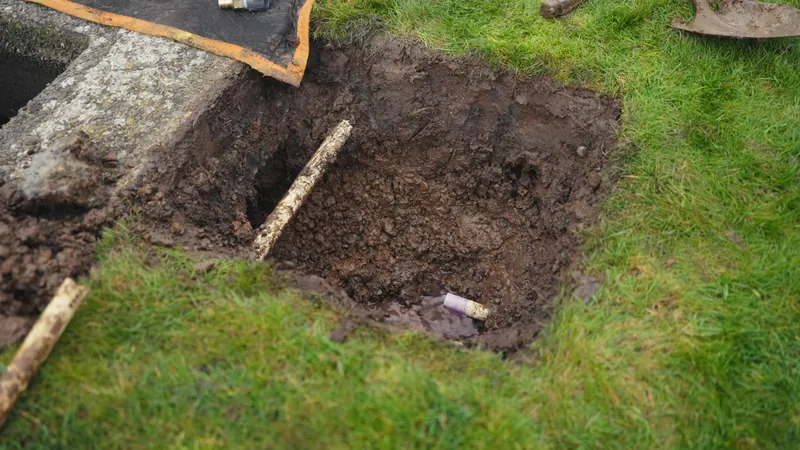 A rectangular hole dug in a grass lawn with loose soil and a stick beside it