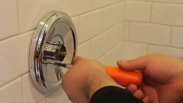 A hand holding an orange screwdriver at the chrome trim plate of a shower valve on a tiled wall