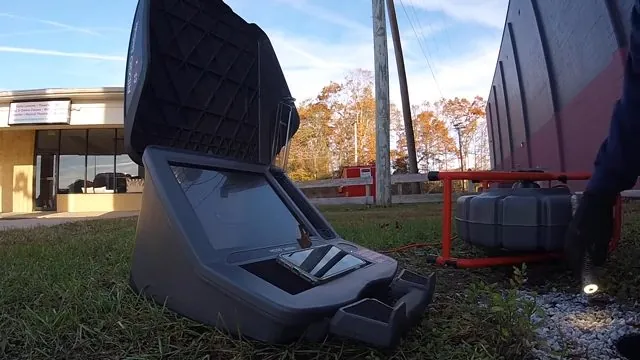 A gray sewer camera control case open on the grass outside a building with autumn trees behind