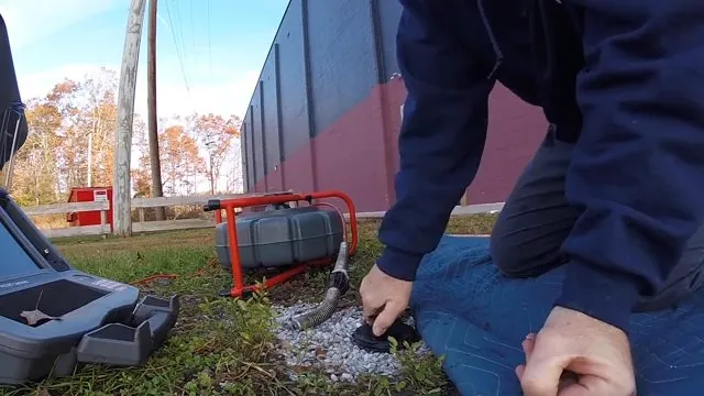 Two people kneeling outdoors at a ground cleanout beside a red camera reel and equipment case