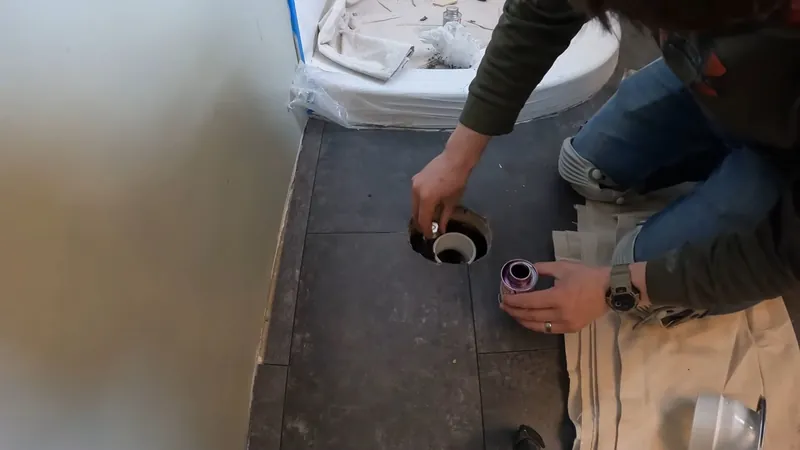 Two people kneeling at a bathroom floor flange with the toilet moved aside
