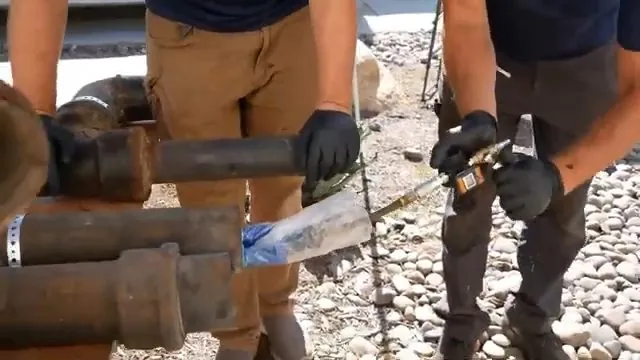 Two workers holding pipe sections and a liner sleeve on a gravel yard