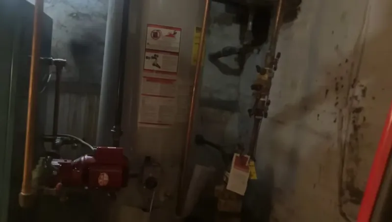 A dimly lit utility room with a water heater, copper and gas pipes and a red valve