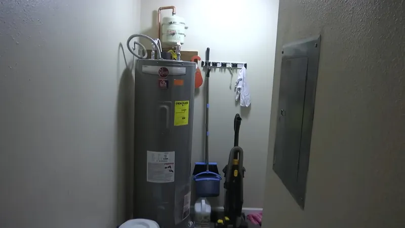A water heater in a utility room with cleaning tools and an electrical panel nearby