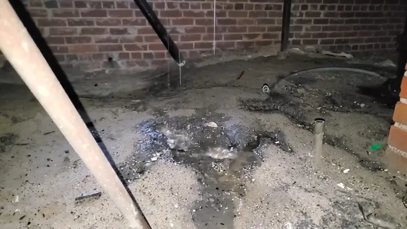 Wet gravel and standing water on the ground under a crawlspace pipe beside a brick wall
