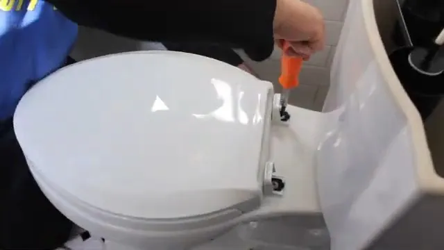 A white toilet with the seat down and a hand holding an orange tool at its base